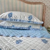 White pillows with blue floral hand block printing on a bed with matching bedspread against a striped headboard. The Charpoy