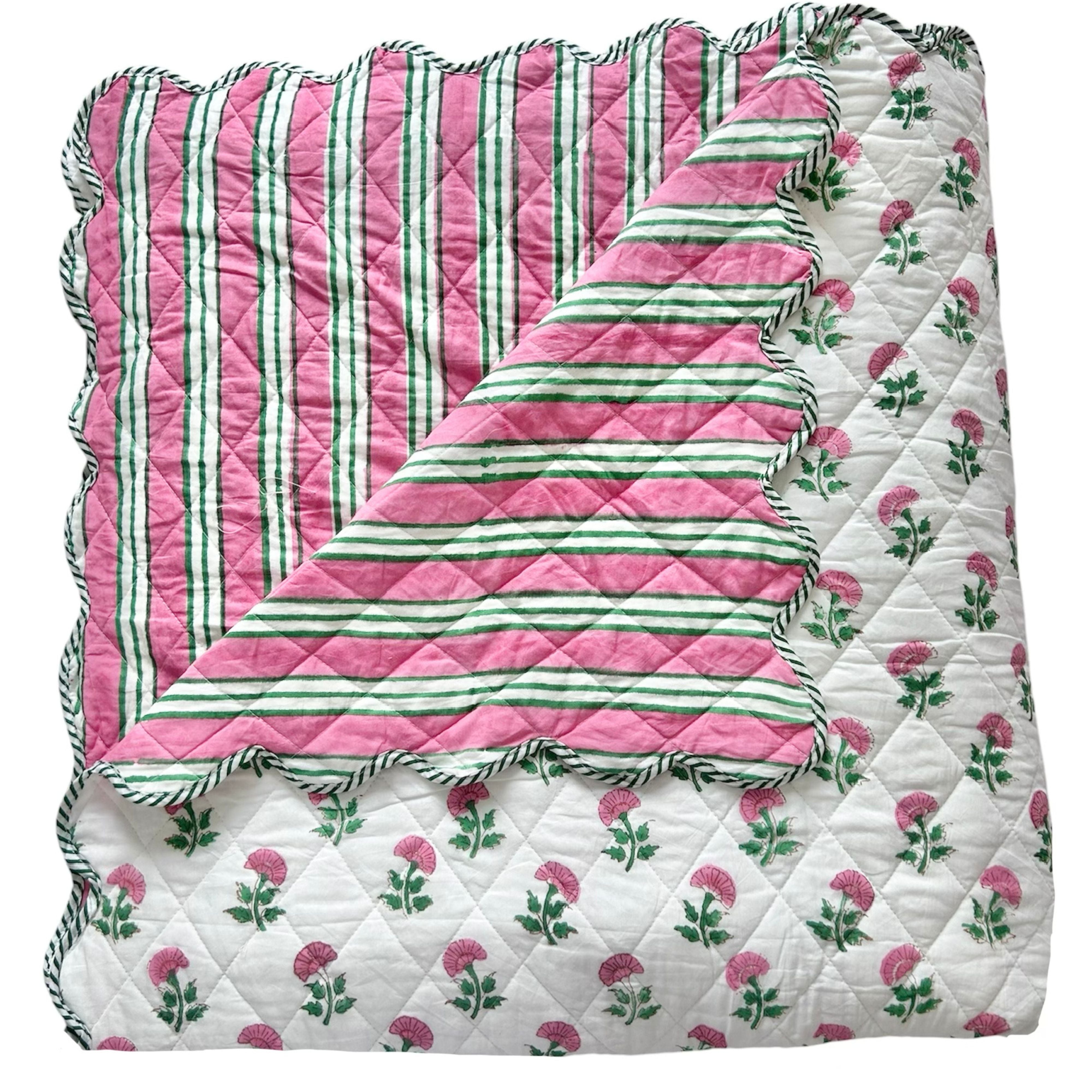 A folded scalloped edge bedspread by The Charpoy with the reverse showing, featuring a repeating pattern of pink flowers and green leaves on a white background. Artisanal handmade in Jaipur. Available in a super king size.