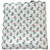 A folded scalloped edge bedspread by The Charpoy, featuring a repeating pattern of pink flowers and green leaves on a white background. Artisanal handmade in Jaipur. Available in a super king size.