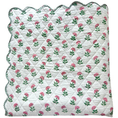 A folded scalloped edge bedspread by The Charpoy, featuring a repeating pattern of pink flowers and green leaves on a white background. Artisanal handmade in Jaipur. Available in a super king size.