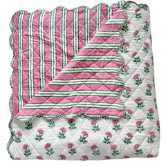 A folded scalloped edge bedspread by The Charpoy with the reverse showing, featuring a repeating pattern of pink flowers and green leaves on a white background. Artisanal handmade in Jaipur. Available in a super king size.