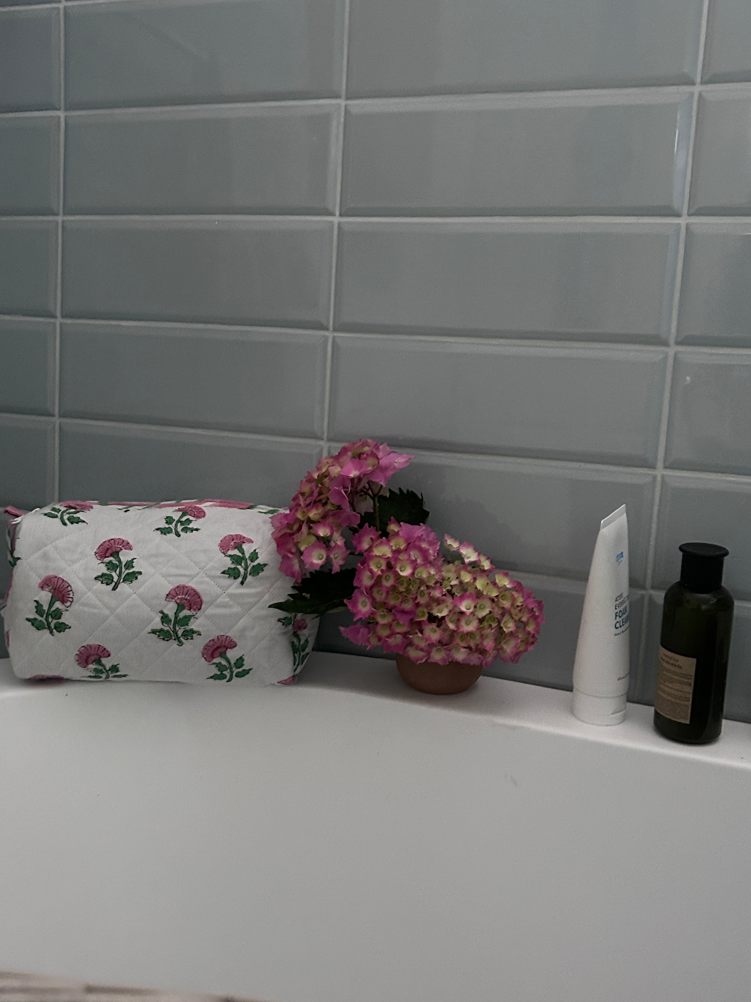 Bathroom setting of a Pink And Green Flower Quilted Wash Bag hand block printed and lightly quilted by artisans in Jaipur by The Charpoy with flowers, and toiletries against a tiled wall.