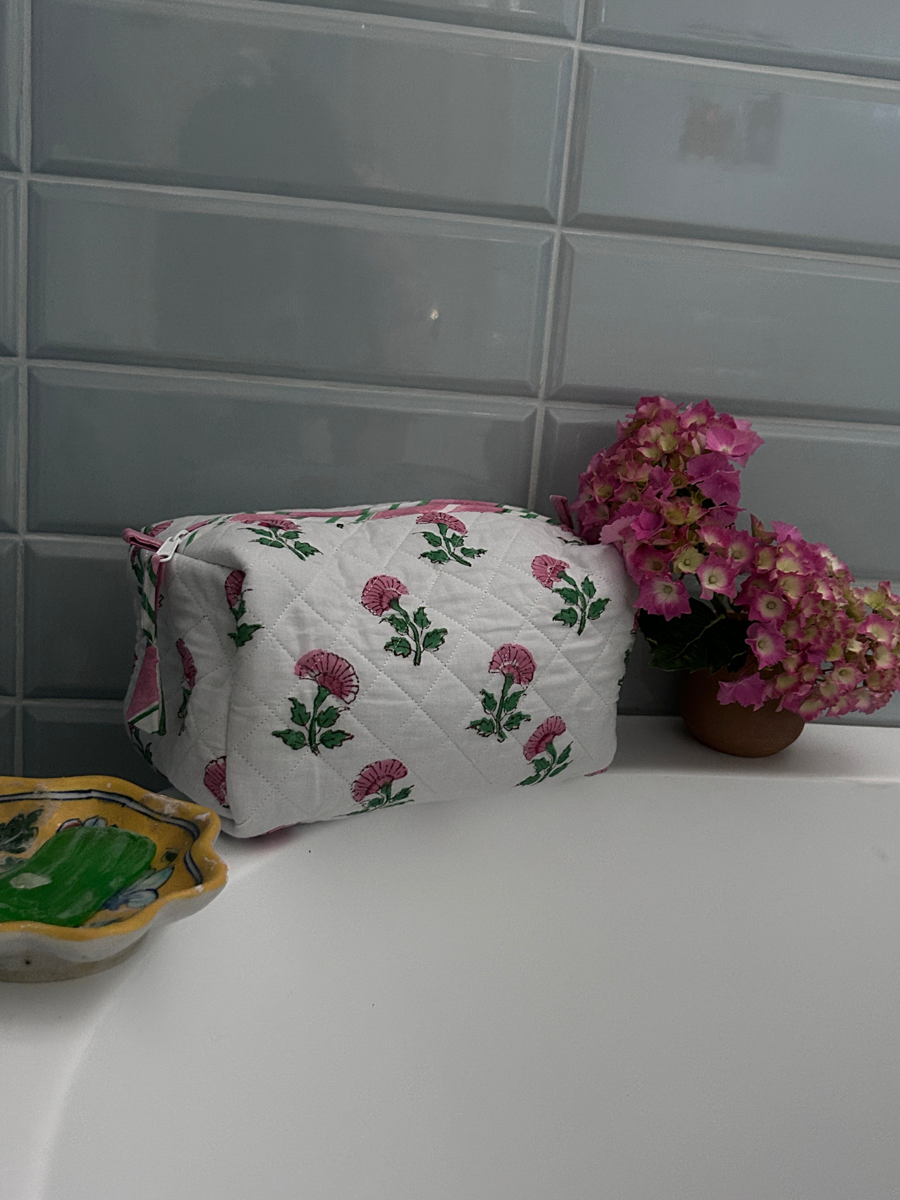 Pink And Green Flower Quilted Wash Bag hand block printed and lightly quilted by artisans in Jaipur by The Charpoy on a bathtub against a tiled wall in bathroom setting.