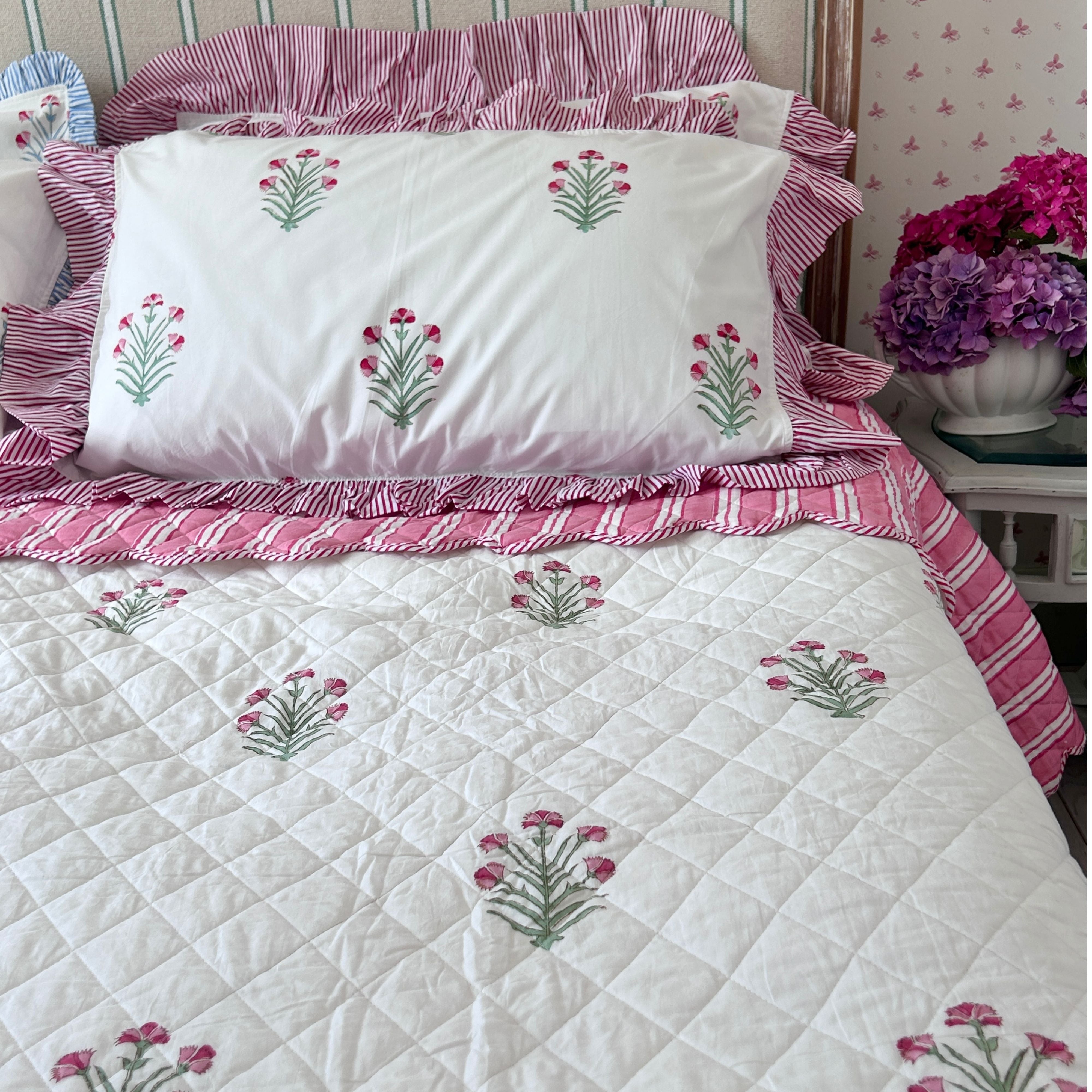 A quilted white bedspread and matching pillowcases decorated with pink and green hand block printed flowers, styled on a bed with pink striped pillows and ruffles in a bedroom setting by The Charpoy