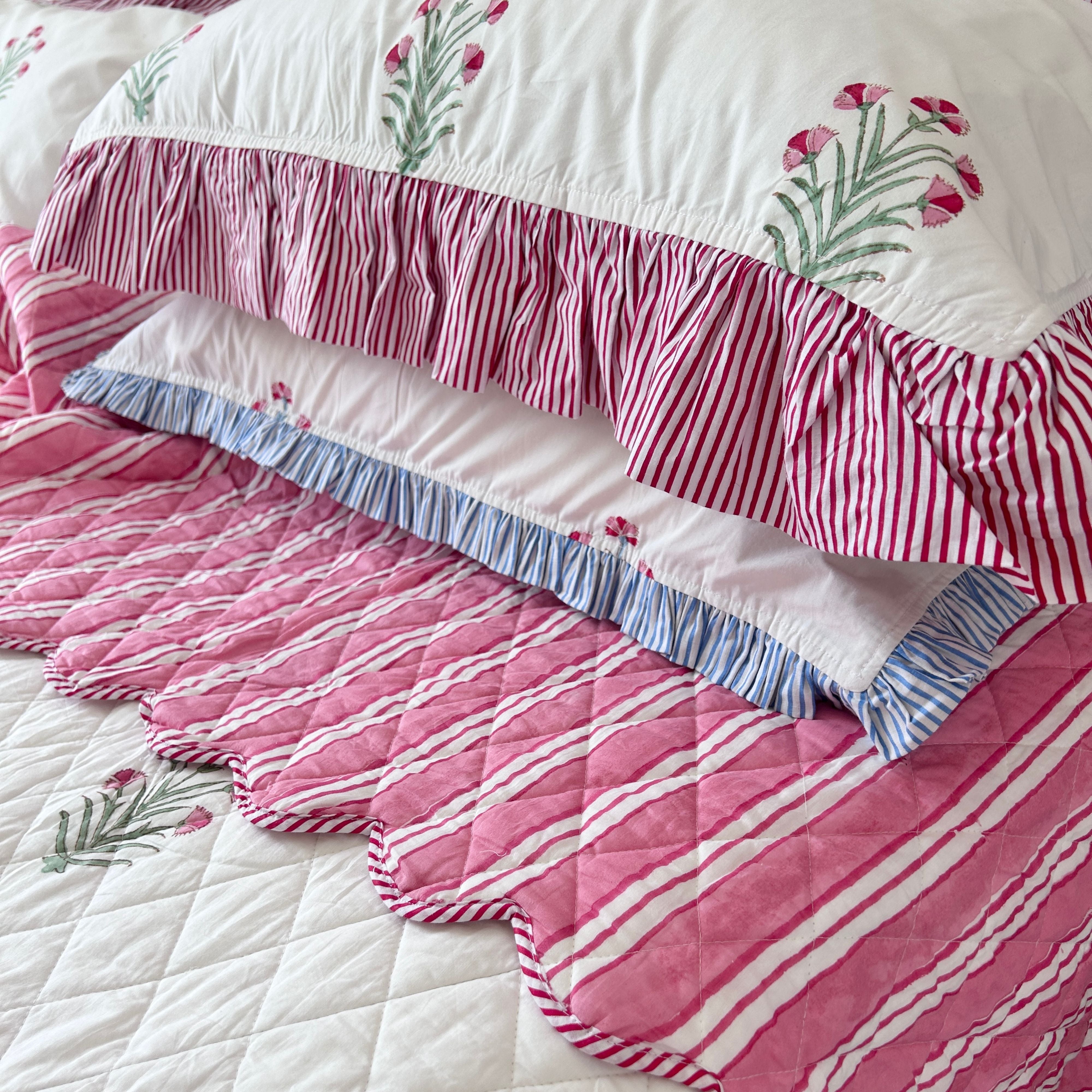 A close-up of a quilted bedding set featuring a pink and white striped quilt with scalloped edges, a matching pink and white striped ruffled bedspread, and white pillows with pink floral and striped ruffles by The Charpoy