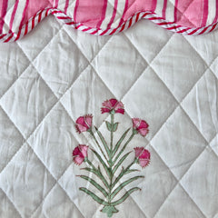 A close-up of a white quilted bedspread with a central hand block printed floral motif in pink and green, and a pink-and-white striped ruffled trim along the top edge by The Charpoy