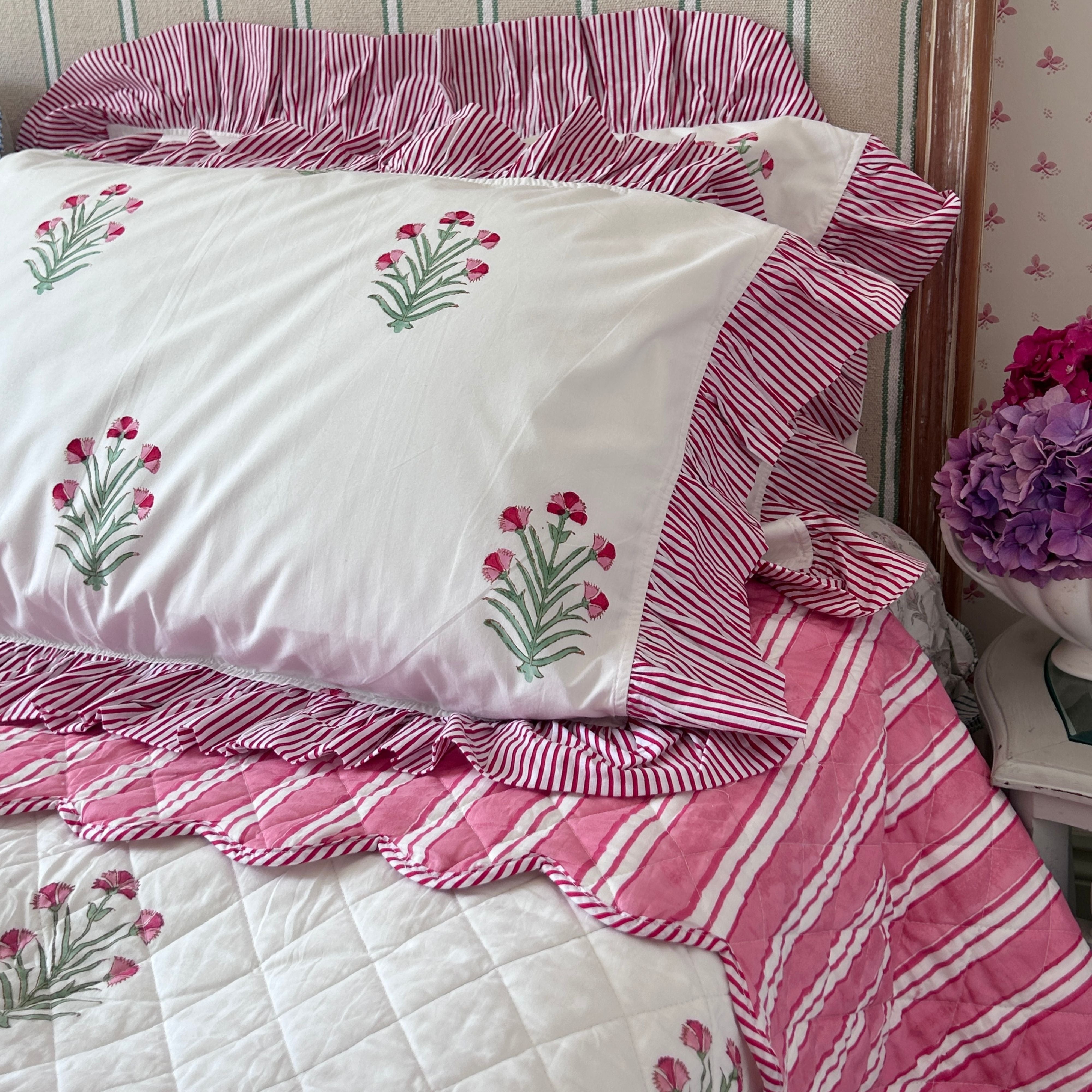 Pink and white striped pillows on a super king bedspread with ruffled edges, featuring a white pillowcase decorated with hand block printed pink flowers and green stems, styled on a bed by The Charpoy