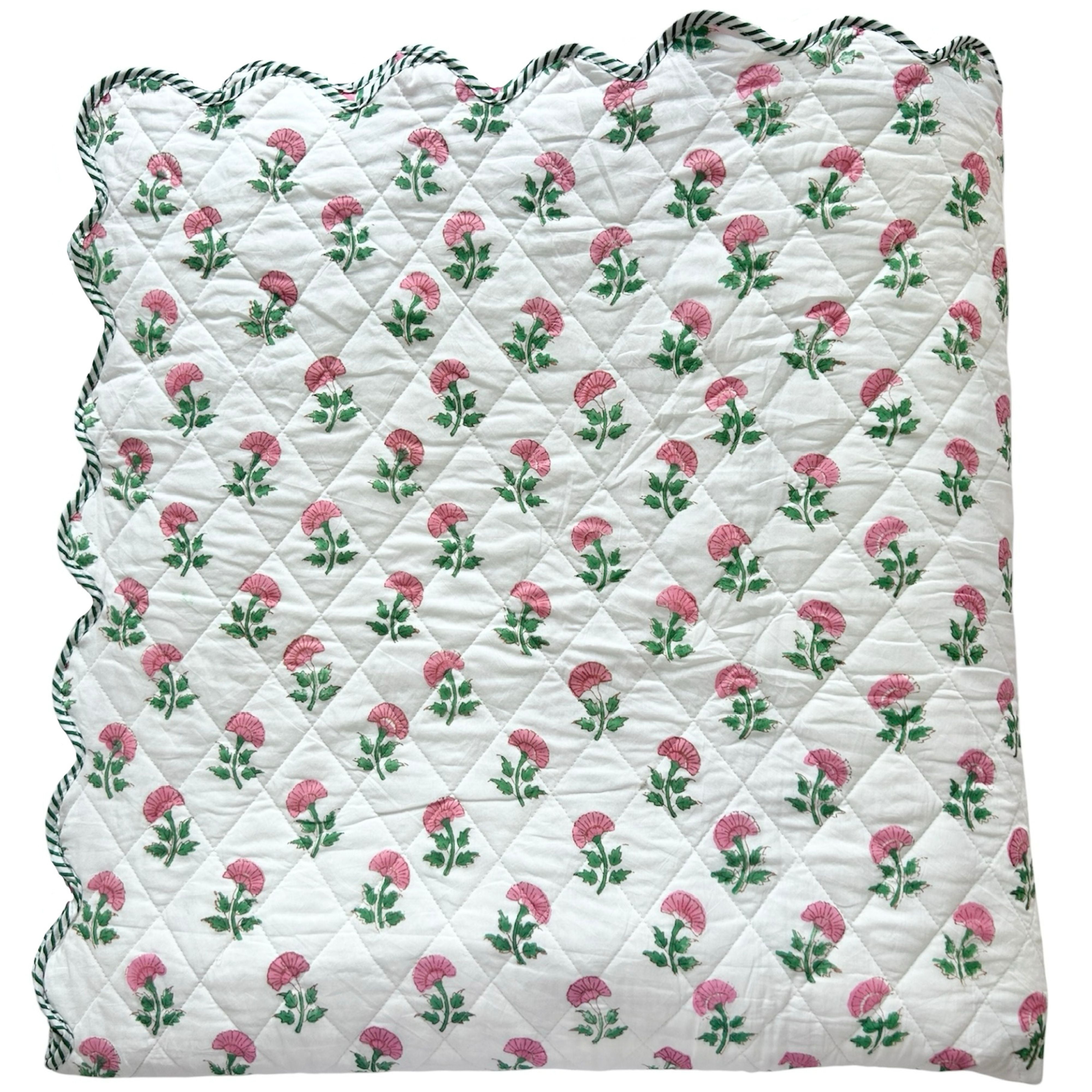 A folded scalloped edge bedspread by The Charpoy, featuring a repeating pattern of pink flowers and green leaves on a white background. Artisanal handmade in Jaipur. Available in a super king size.