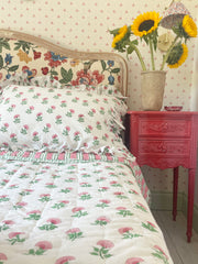 A bed with ruffled pillow covers featuring a pink carnation design, placed in a bedroom setting with a red nightstand and a vase of sunflowers on the side.