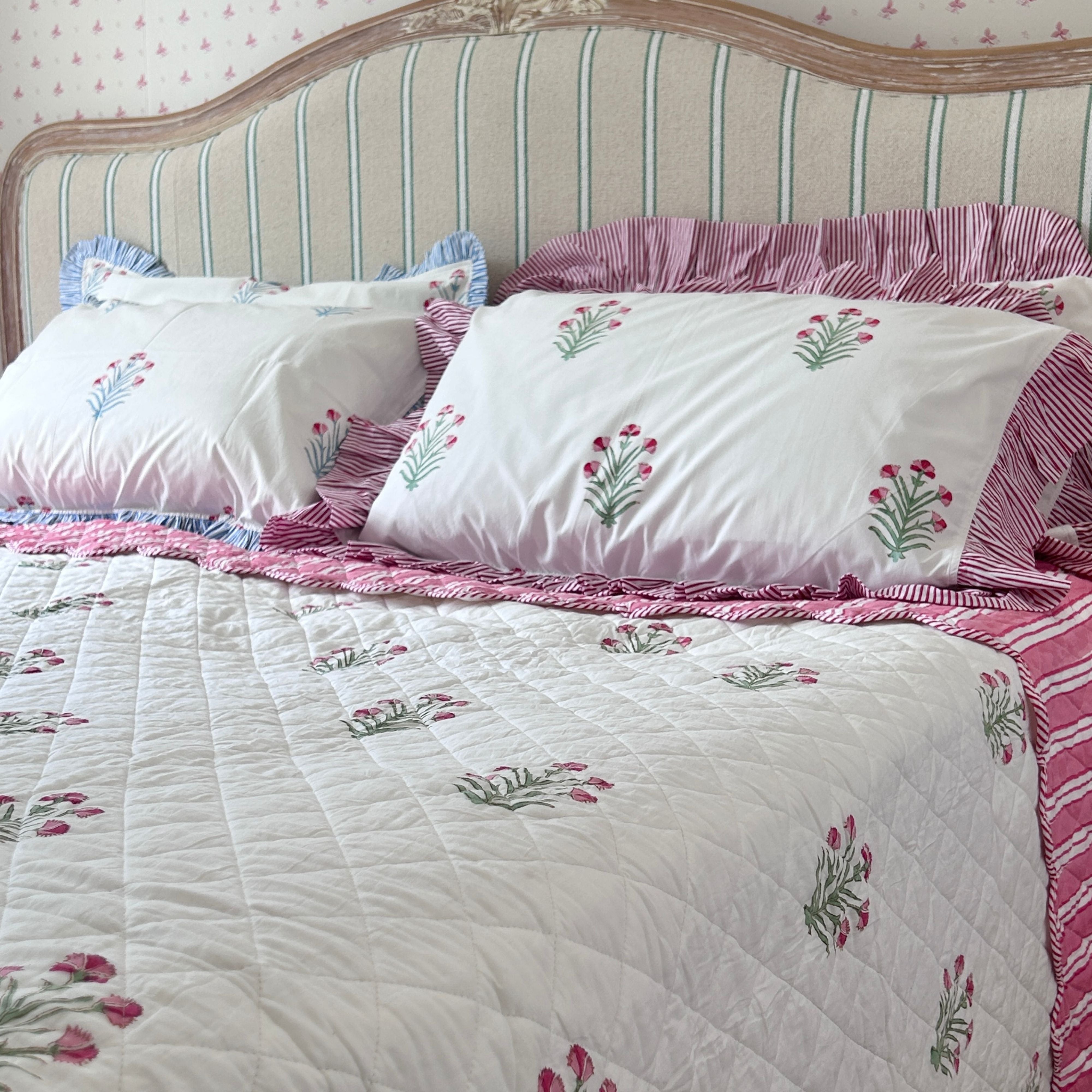 A bed dressed in pink and green buta  done with hand block printing, layered  and matched with pink flowers pillow covers set against a cream and blue striped headboard by The Charpoy