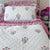 A quilted white bedspread and matching pillowcases decorated with pink and green hand block printed flowers, styled on a bed with pink striped pillows and ruffles in a bedroom setting by The Charpoy