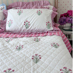 A quilted white bedspread and matching pillowcases decorated with pink and green hand block printed flowers, styled on a bed with pink striped pillows and ruffles in a bedroom setting by The Charpoy
