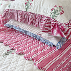 A close-up of a quilted bedding set featuring a pink and white striped quilt with scalloped edges, a matching pink and white striped ruffled bedspread, and white pillows with pink floral and striped ruffles by The Charpoy