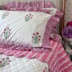 Pink and white striped pillows on a super king bedspread with ruffled edges, featuring a white pillowcase decorated with hand block printed pink flowers and green stems, styled on a bed by The Charpoy